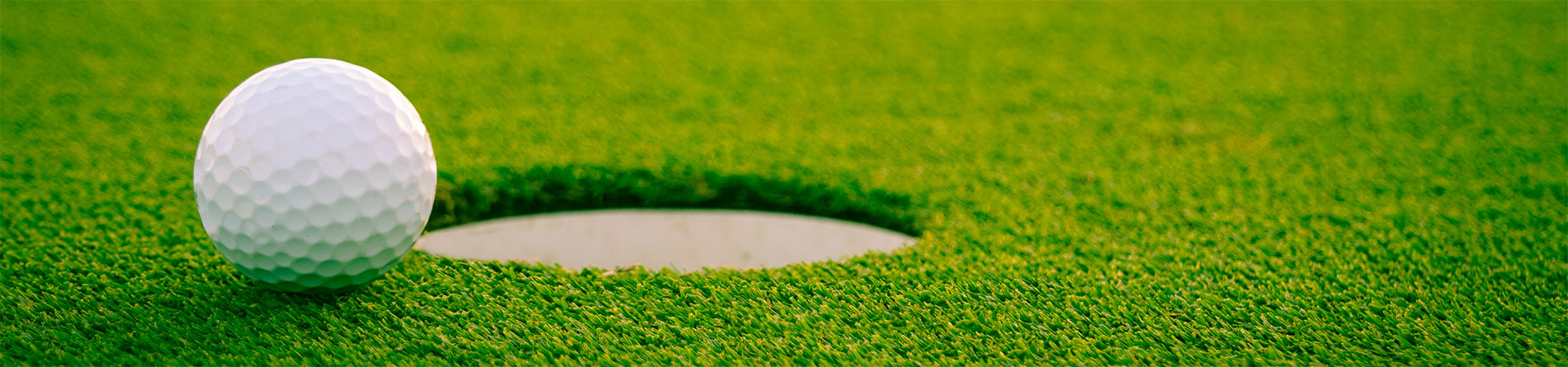 A close-up view of a textured white golf ball just inches from the hole on a vibrant green putting green.